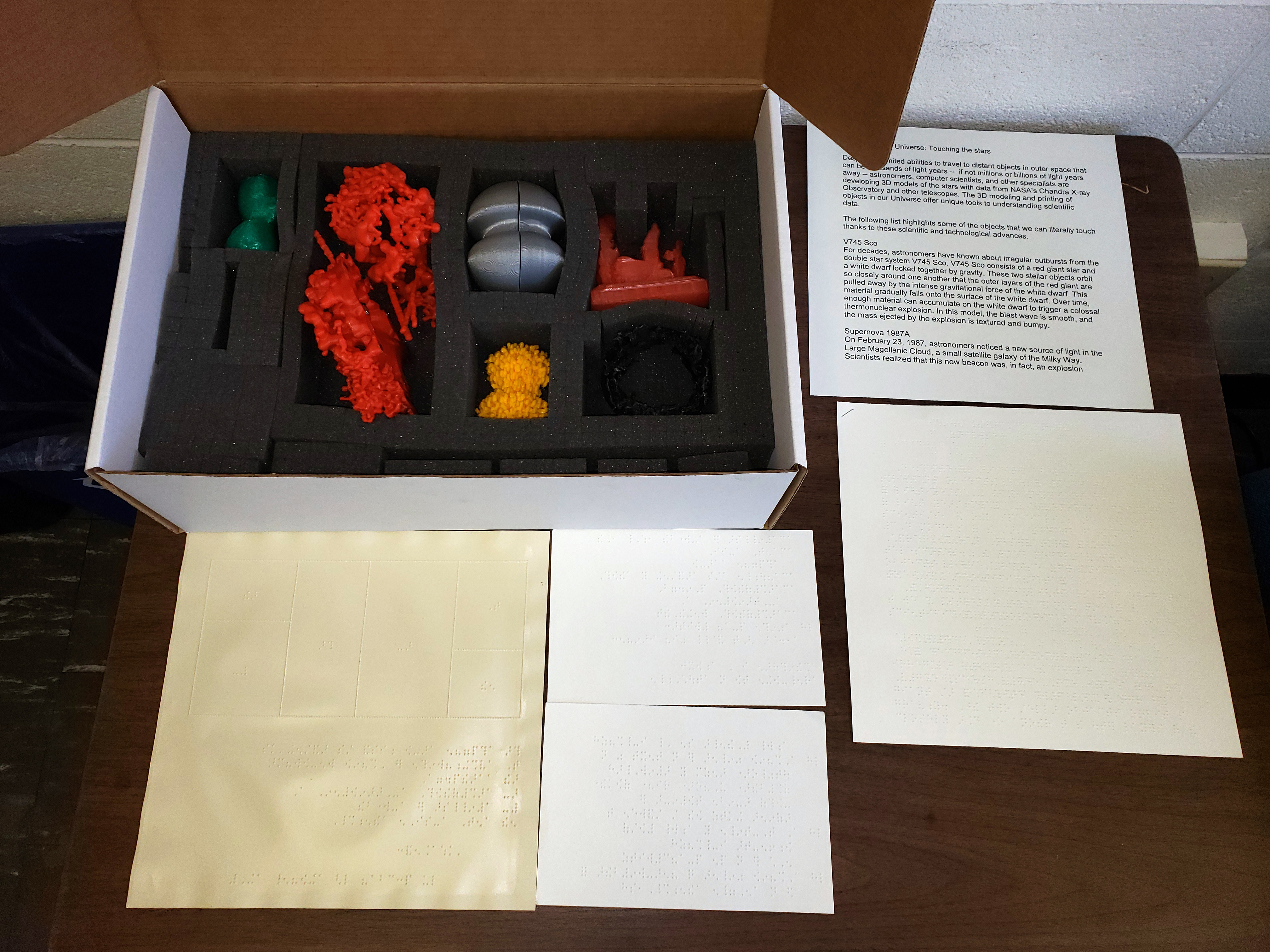 Photo of a 12-inch box open to display a collection of small 3D printed astronomy models. Descriptions of models in braille lay in front of the box.